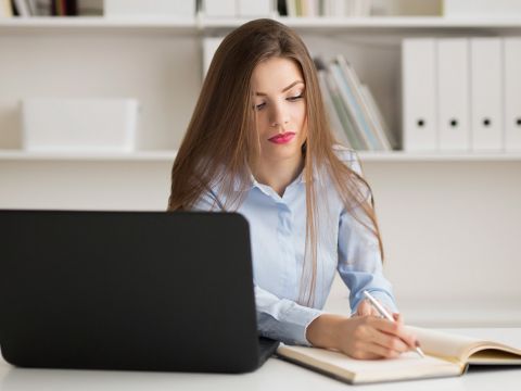 Frau sitzt vor Laptop, notiert sich Dinge in ein Notizbuch. Im Hintergrund ist ein Regal mit Ordnern und Dokumenten.