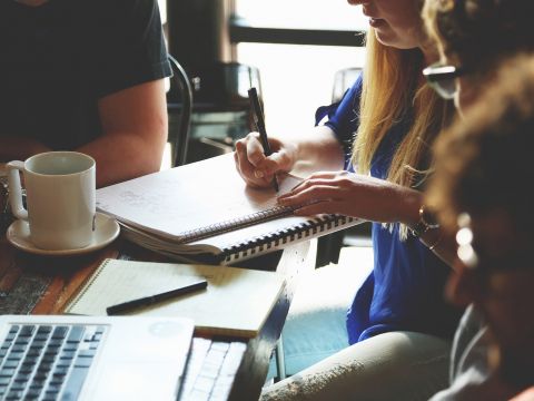 Es findet ein Meeting von mindestens drei Personen statt. Ein Laptop und ein Notizblock liegen auf dem Tisch und eine Frau schreibt etwas in ein Notizbuch.